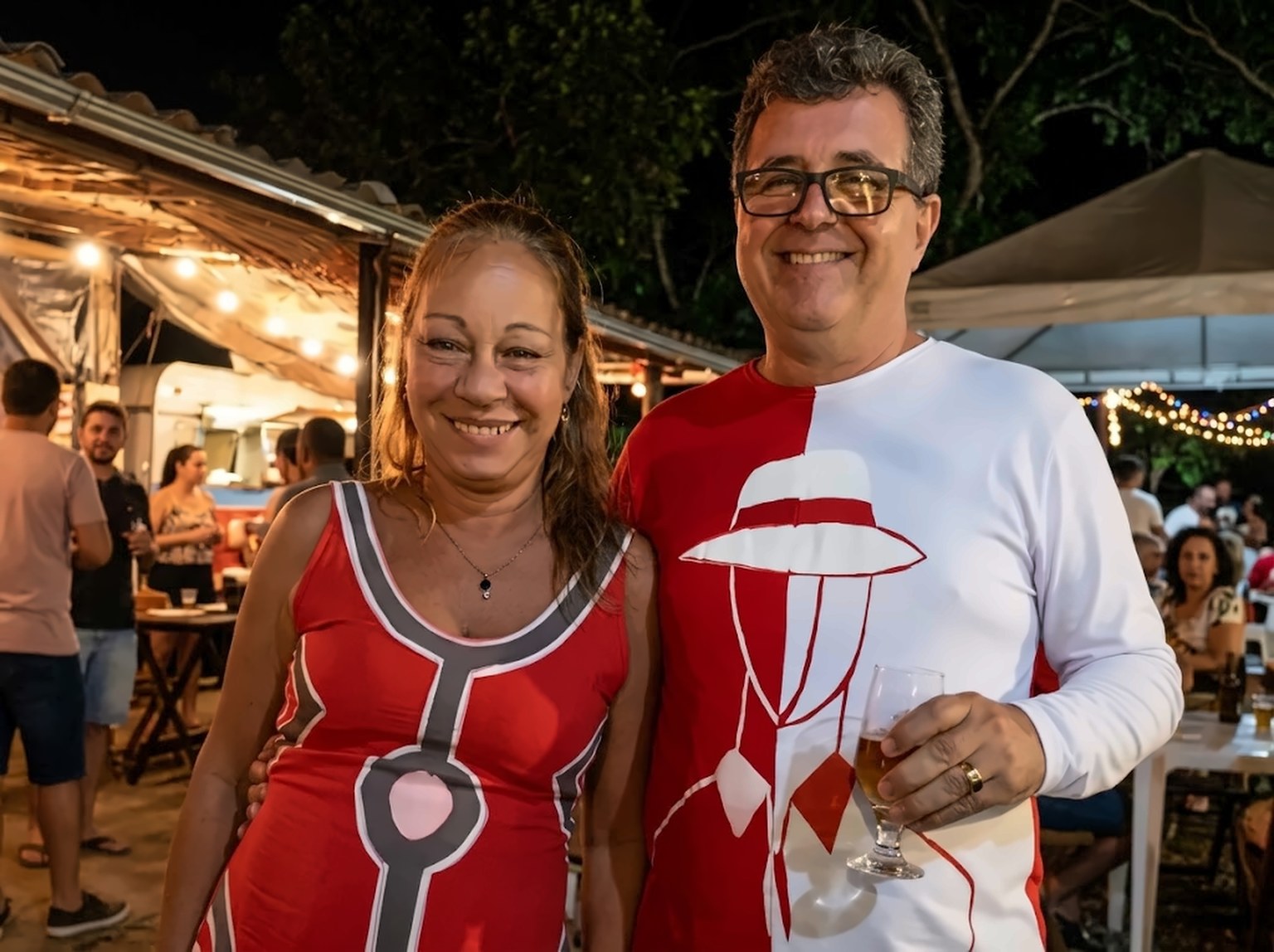 Rosa Tibau e Rogério Pinto sorrindo em um evento, ela com vestido vermelho e ele com camiseta vermelha e branca, ambos pintados à mão.