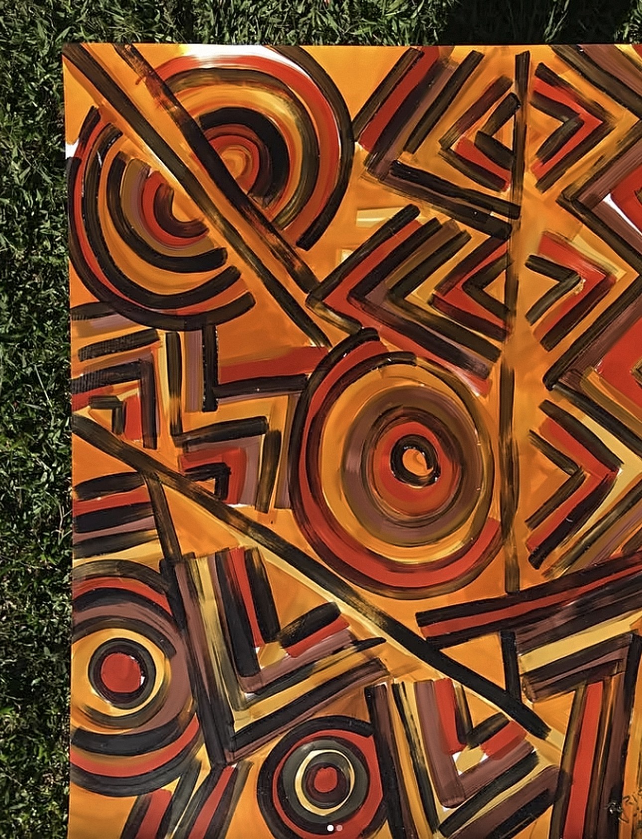 Detalhe de um quadro em laranja, preto e vermelho, com círculos concêntricos, zigue-zagues e linhas diagonais, sobre o gramado.