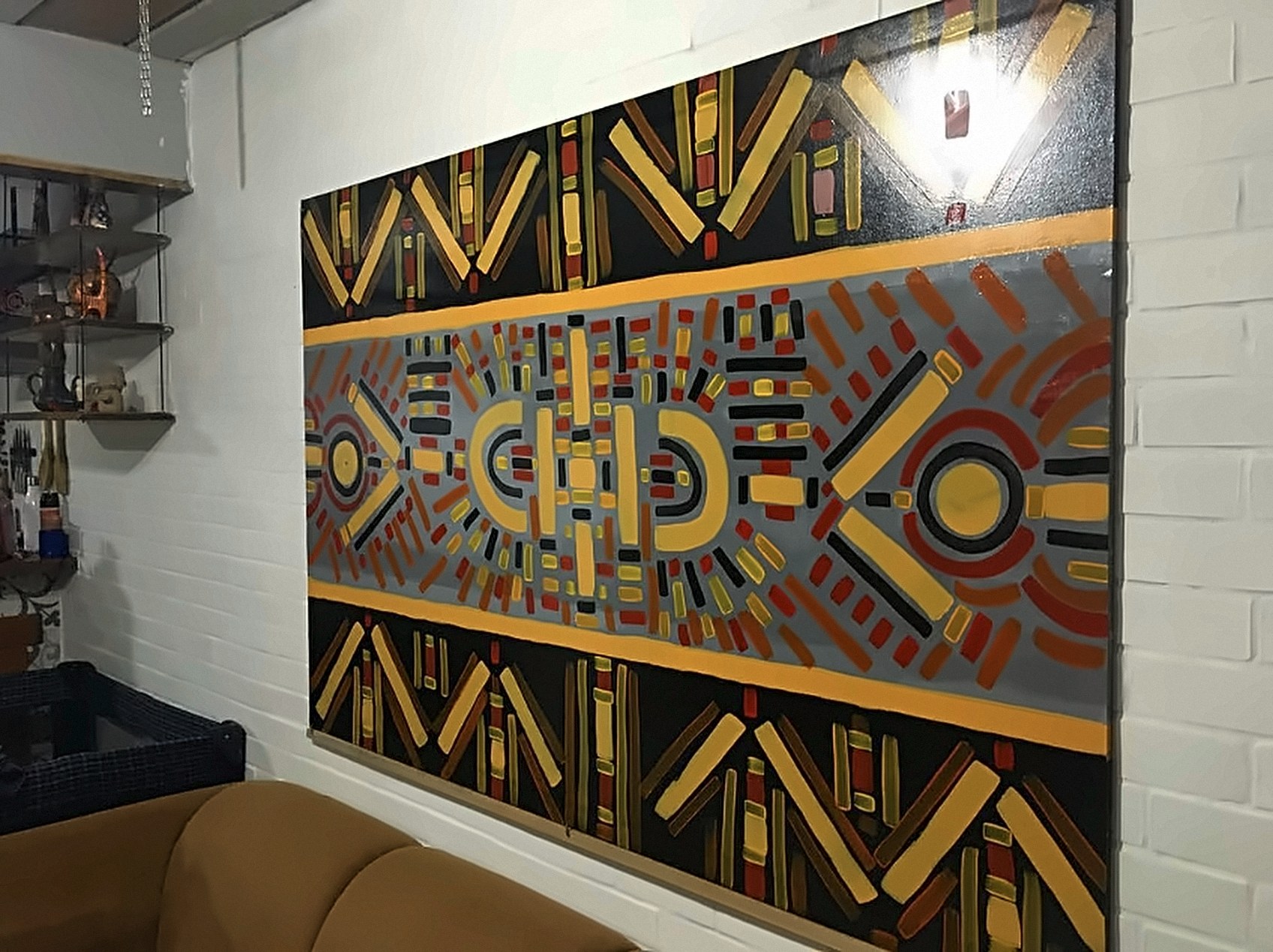 Quadro grande pendurado em parede de tijolos brancos, com faixa cinza central de formas em vermelho, amarelo e preto entre faixas pretas com listras amarelas.