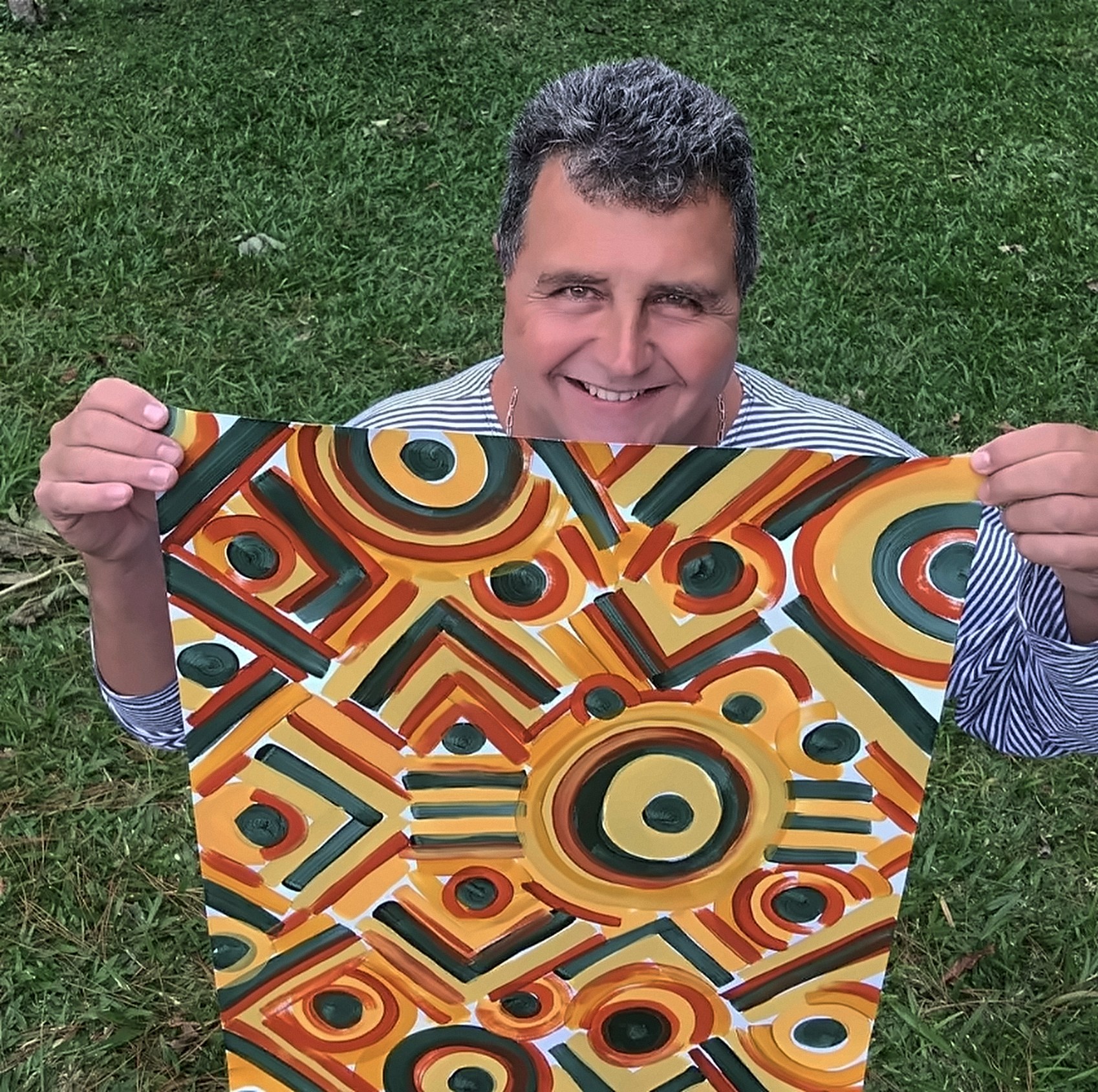 Homem sorrindo, ao ar livre sobre o gramado, segurando uma pintura em tons de laranja, verde-escuro e vermelho, com círculos concêntricos e linhas em zigue-zague.