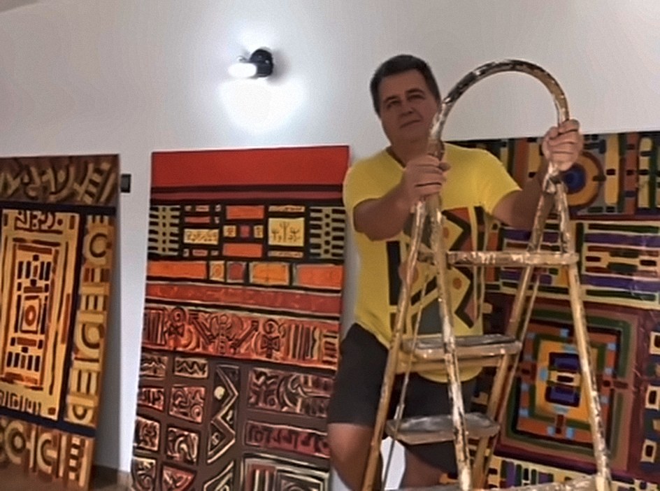 Homem de camiseta amarela segurando uma escada de pintor, em uma sala com telas grandes pintadas em vermelho, laranja e amarelo ao fundo.