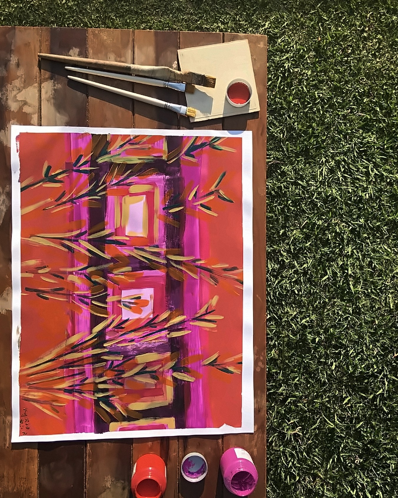 Pintura em laranja, rosa e roxo com folhas finas, sobre uma mesa de madeira no gramado, com três pincéis e potes de tinta vermelha, roxa e rosa.
