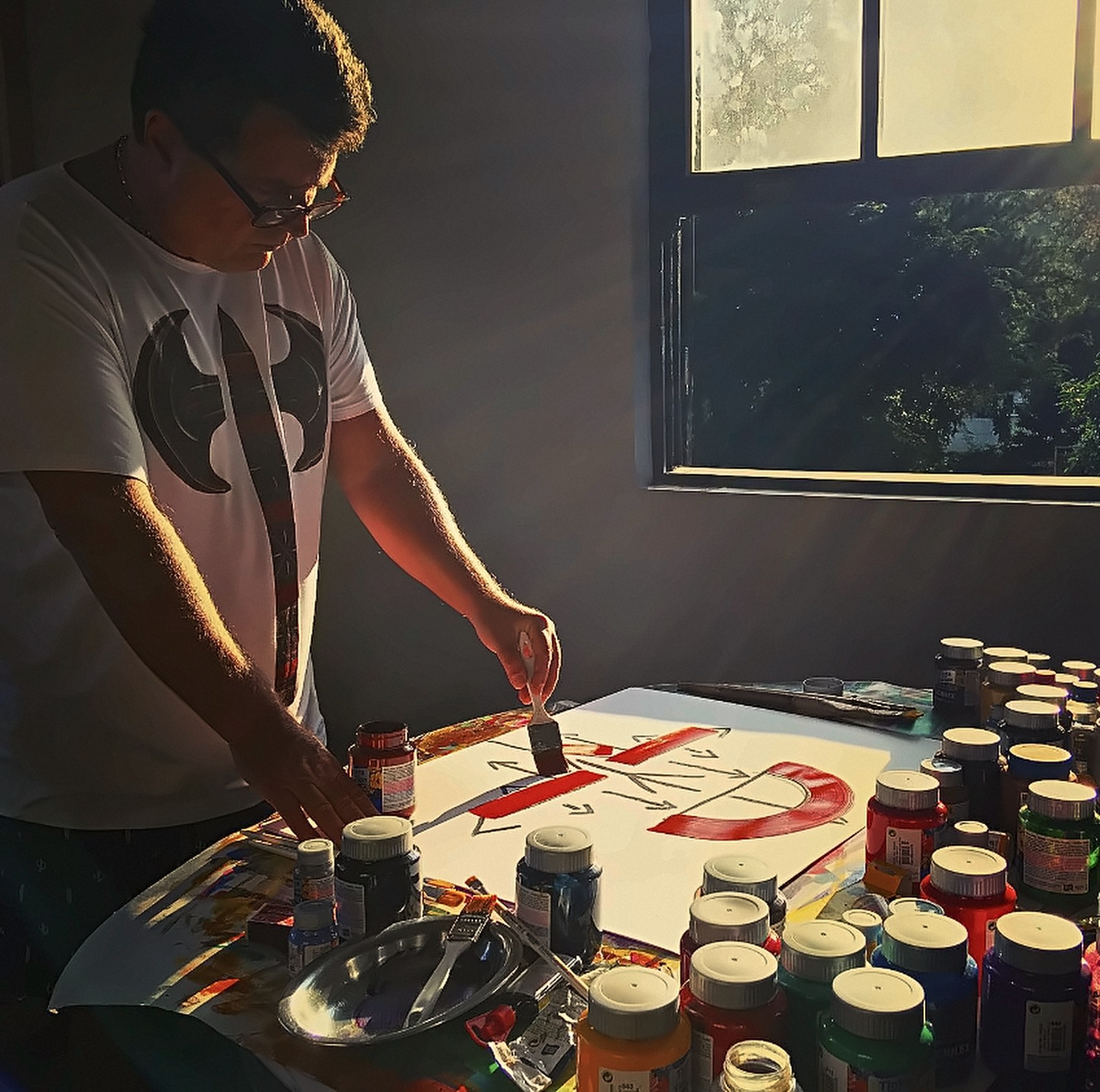 Homem de óculos pintando com pincel sobre uma folha branca, em uma mesa cheia de potes de tinta coloridos, ao lado de uma janela com luz forte entrando.