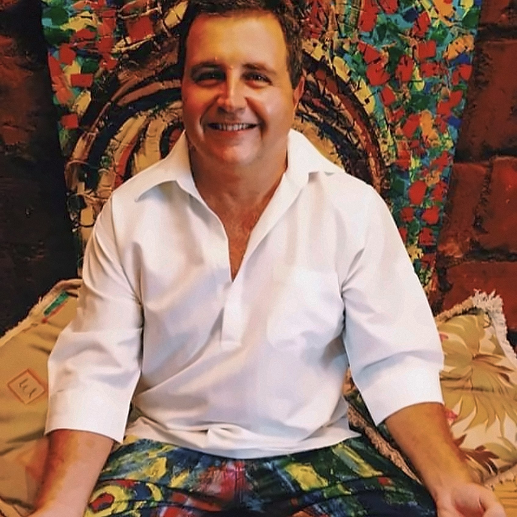 Homem sorrindo, de camisa branca, sentado em frente a um quadro de fragmentos coloridos em vermelho, verde, azul e amarelo, sobre almofadas estampadas.