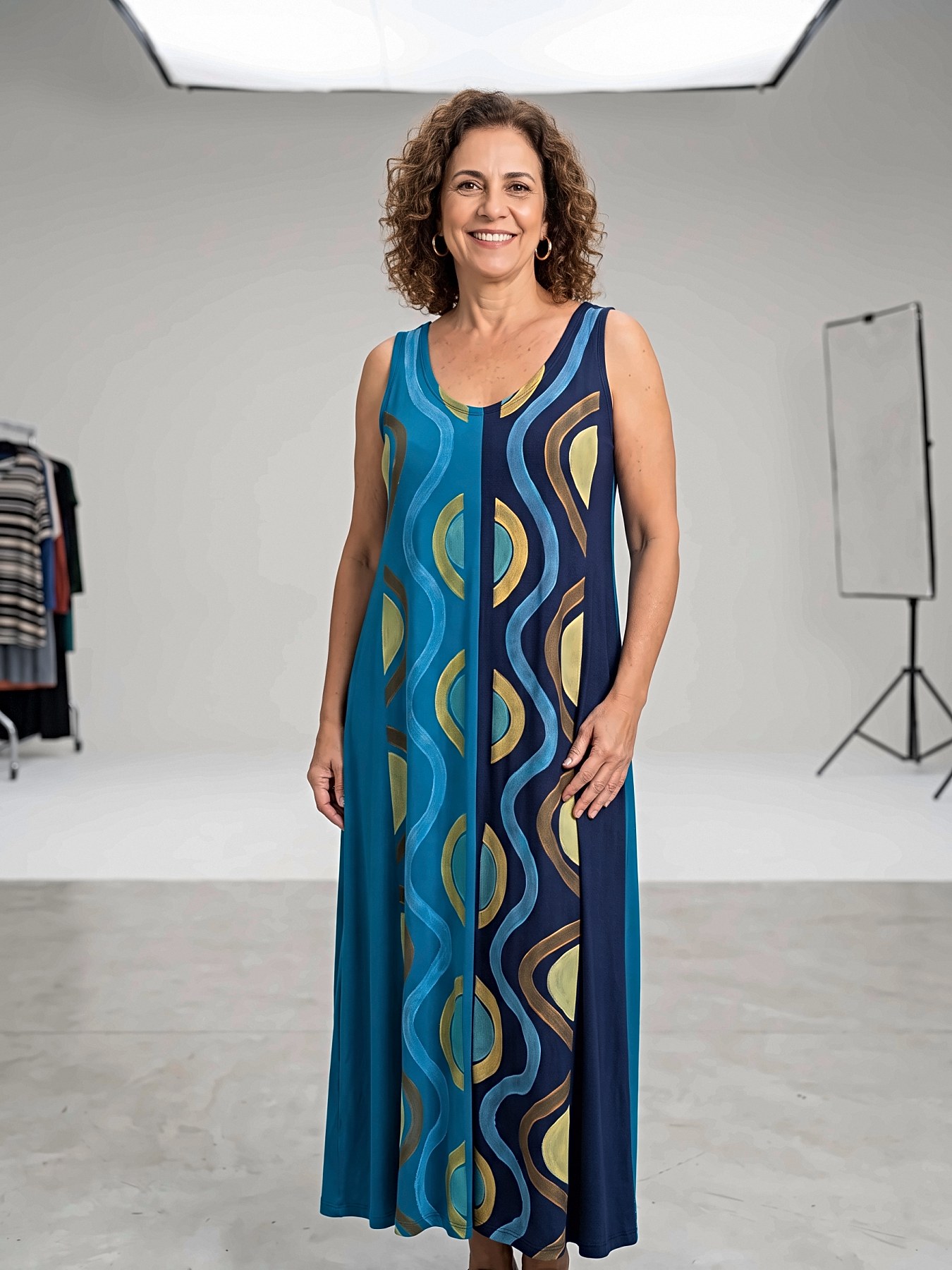 Mulher sorrindo, de pé em estúdio, vestindo vestido longo sem manga dividido em azul-petróleo e azul-marinho, com ondas e formas pintadas à mão em azul claro, dourado e marrom.