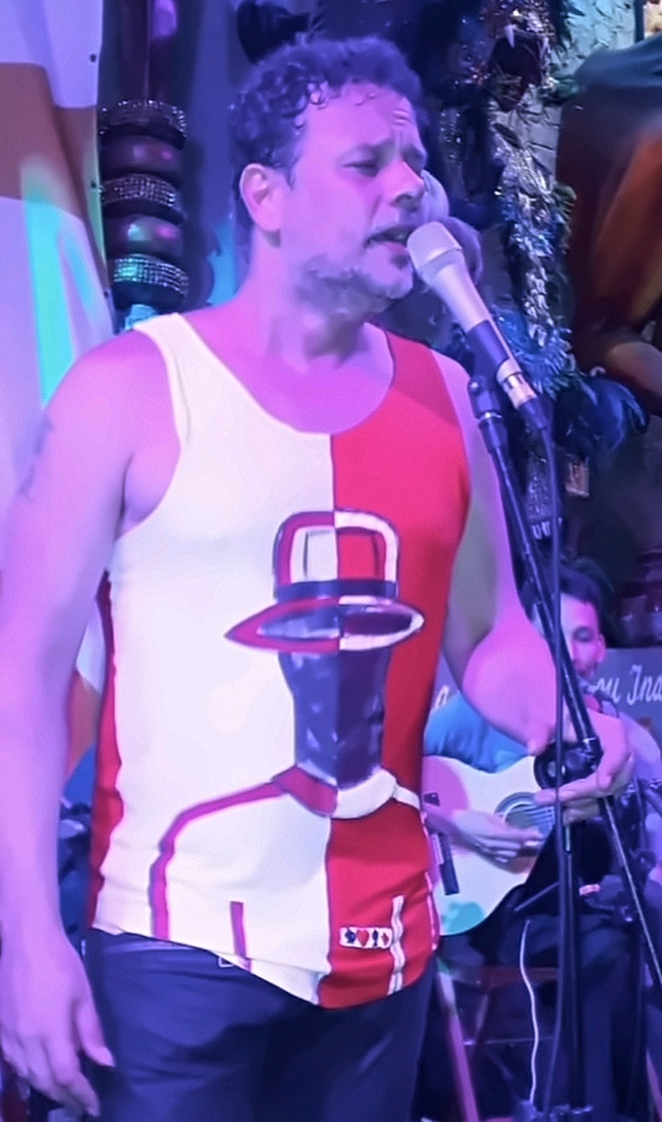 Toninho Geraes cantando ao microfone, vestindo regata dividida em branco e vermelho, com o desenho pintado de um homem de chapéu.