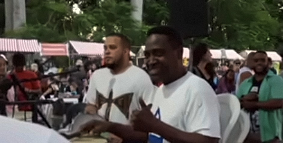 Artistas em um evento ao ar livre, vestindo camisetas brancas com estampas da Tribau, um deles sorrindo e batendo palmas.