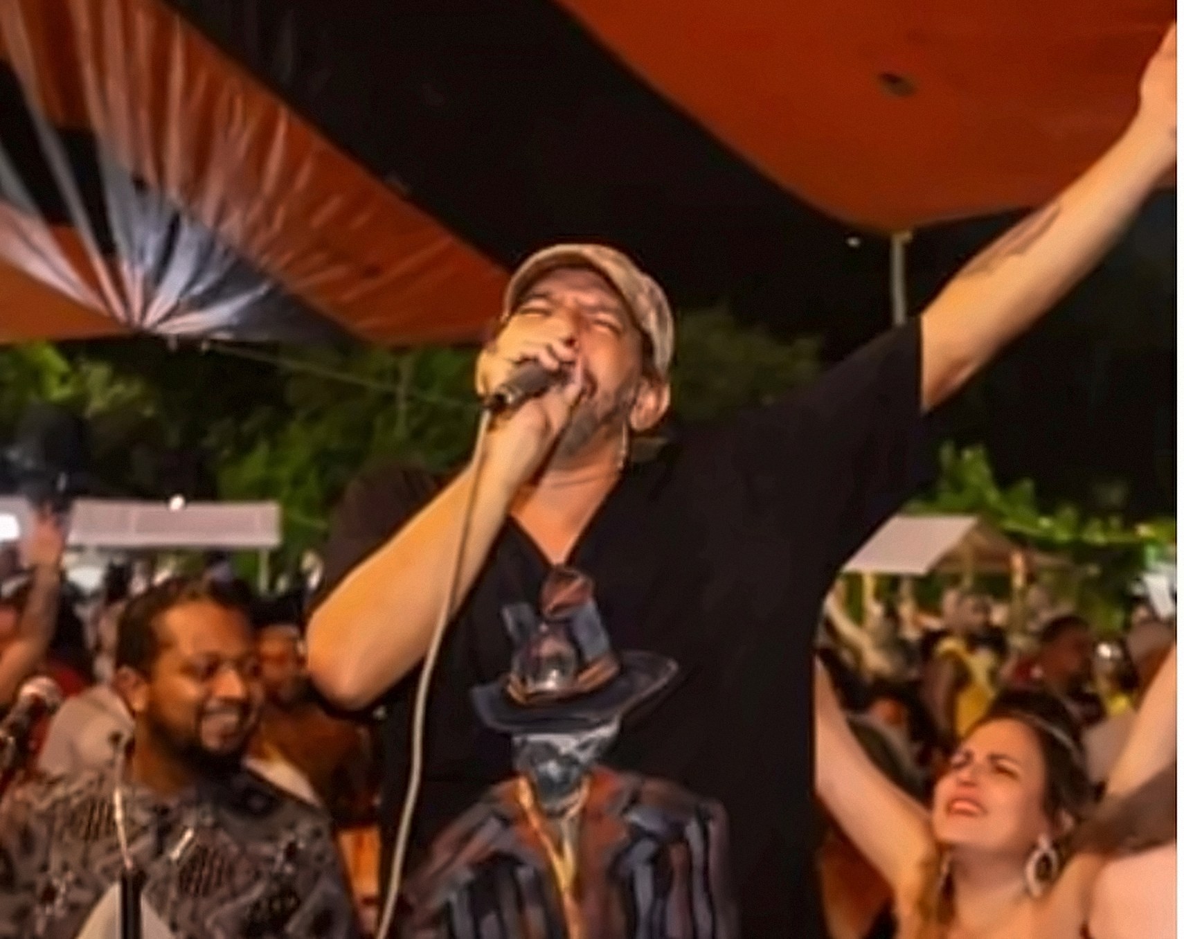 Artista cantando no palco ao ar livre, de braço levantado, vestindo camiseta preta com o desenho pintado de um homem de chapéu e paletó listrado.