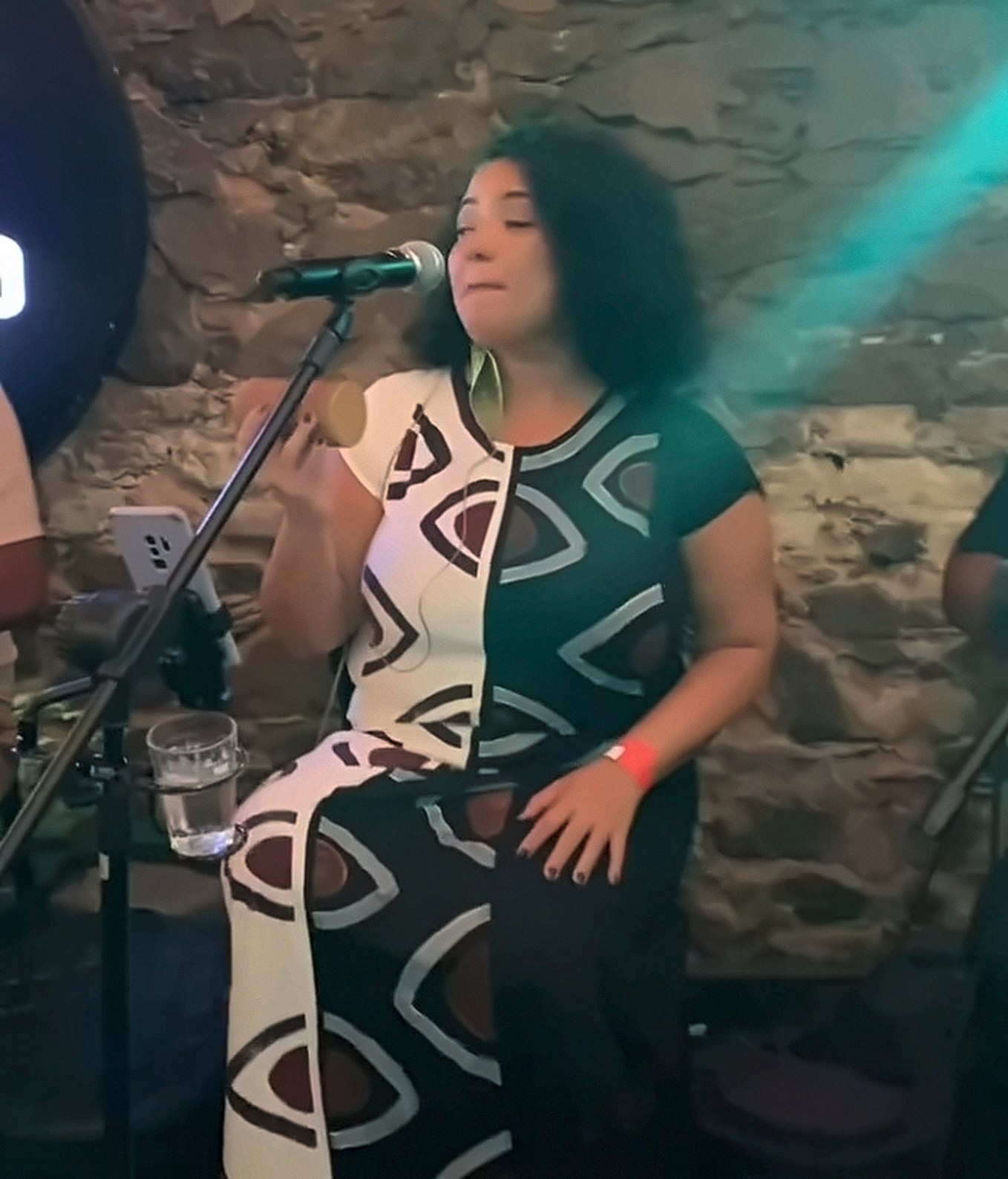 Cantora ao microfone, sentada diante de uma parede de pedra, vestindo vestido dividido em branco e preto com olhos pintados à mão.