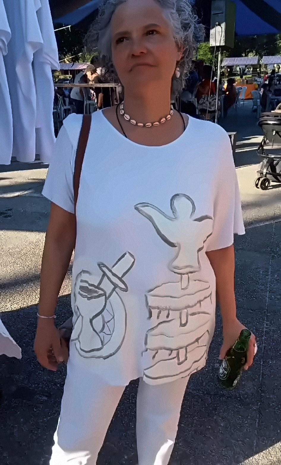 Mulher de cabelo grisalho em um evento ao ar livre, vestindo camiseta branca ampla com desenhos pintados à mão em cinza.