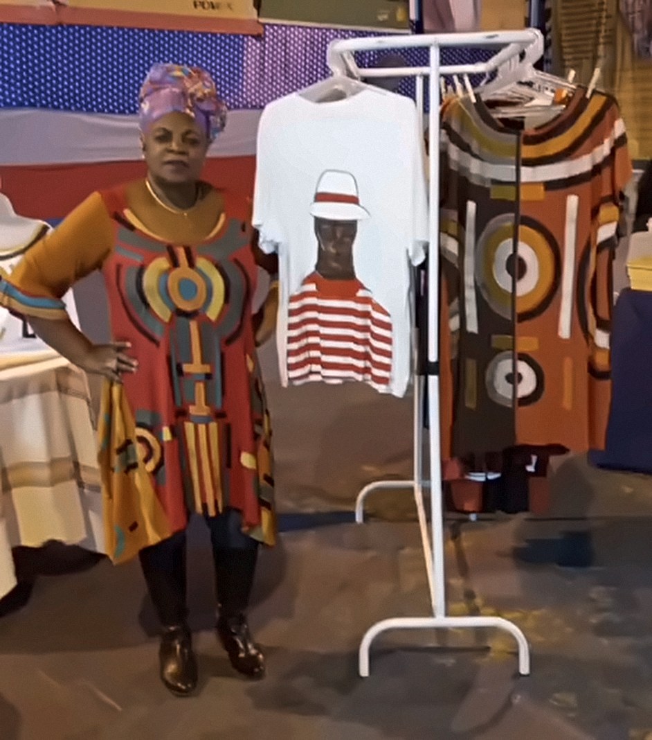 Mulher de turbante colorido e túnica pintada em vermelho, laranja e amarelo, ao lado de uma arara com camisetas e vestidos pintados à mão.