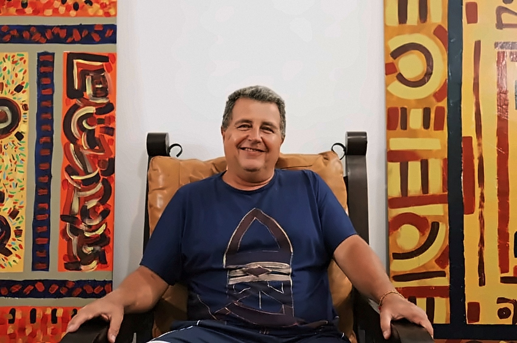 Homem sorrindo, sentado em uma poltrona de madeira entre dois quadros grandes, vestindo camiseta azul-marinho com um desenho pintado à mão.